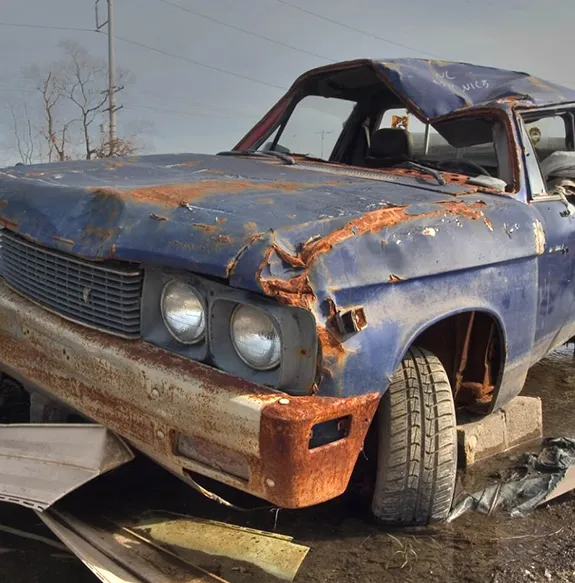 A heavily rusted, abandoned car with a crushed roof.