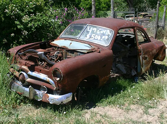 A heavily rusted, abandoned 1949 Ford coupe in a junkyard.